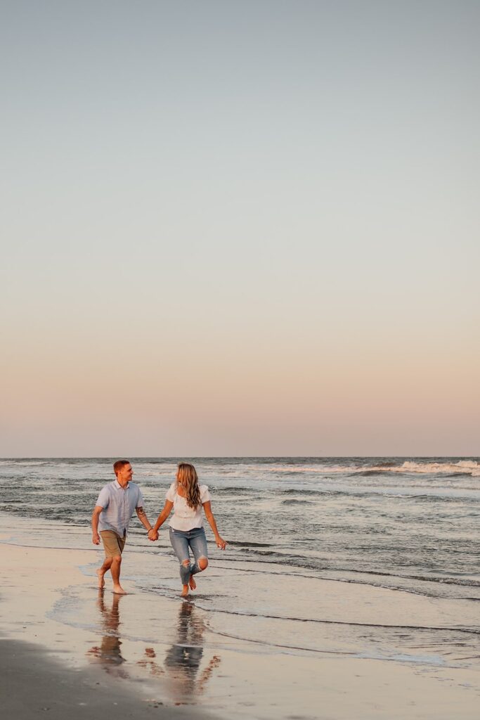 Couples holds hands while running across the beach during their engagement session in Folly Beach, South Carolina
