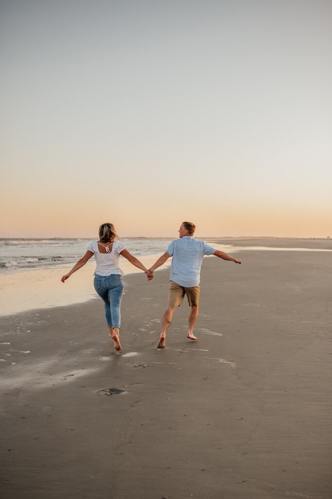 Couple holds hands while running across the sand during their beach enegagement session