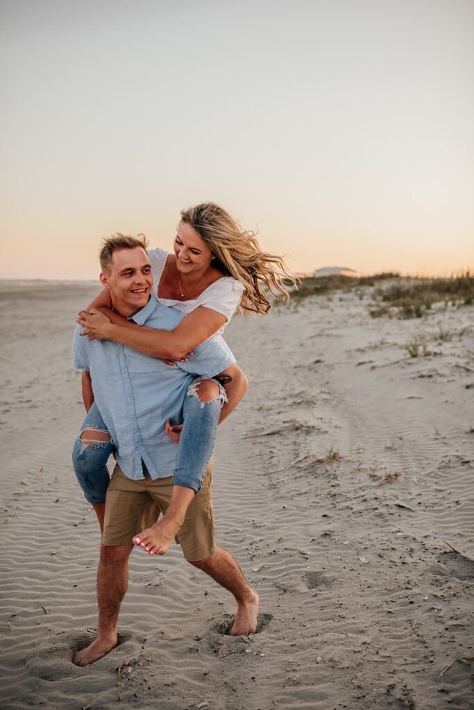 Man carrying woman piggy-back style on the beach during their Charleston engagement photos