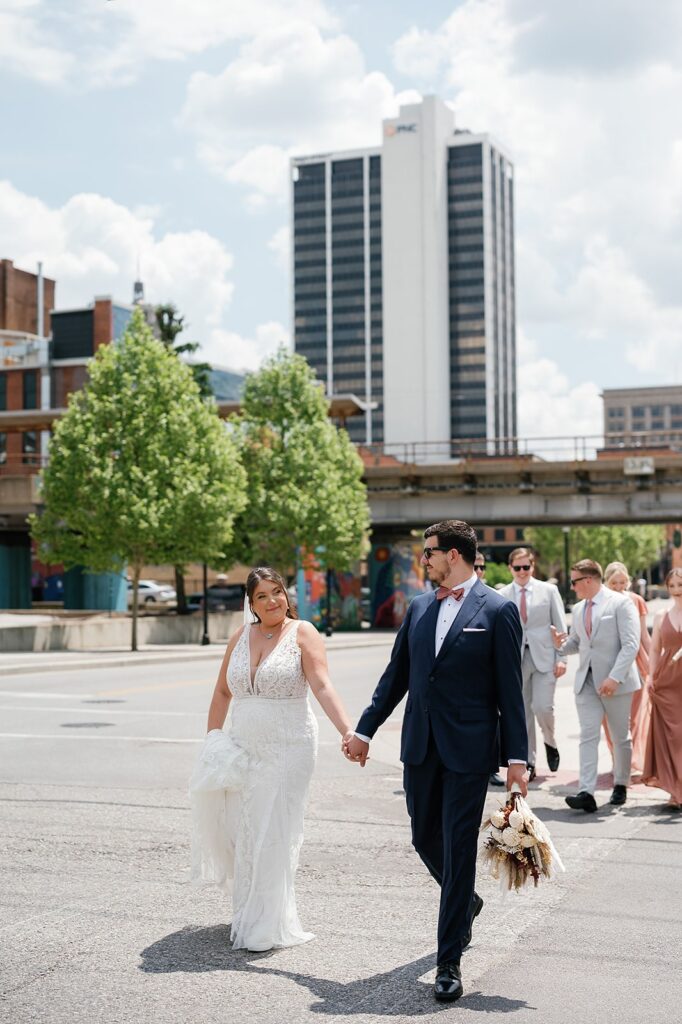 Bride and groom hold hands as they walk with their wedding party through the streets of downtown Fort Wayne