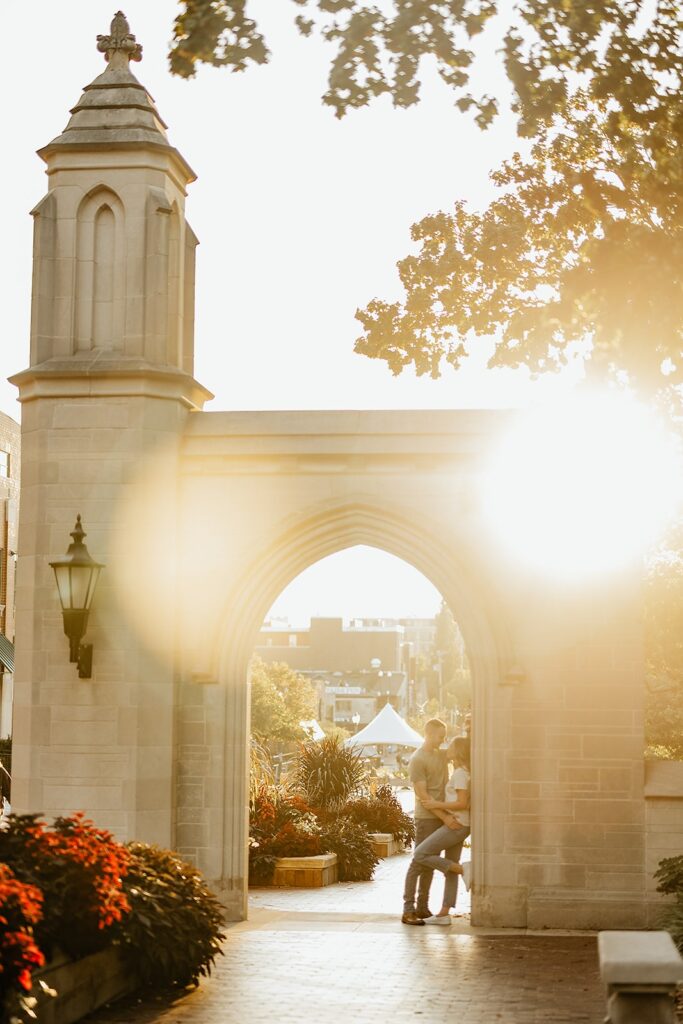 Man and woman stand under an arch at sunset during their engagement session at Indiana University
