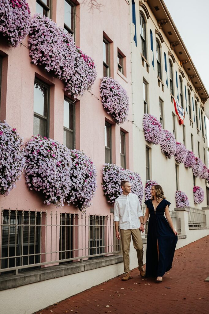 Man and woman hold hands while walking down a street with pink flower boxes in the building windows
