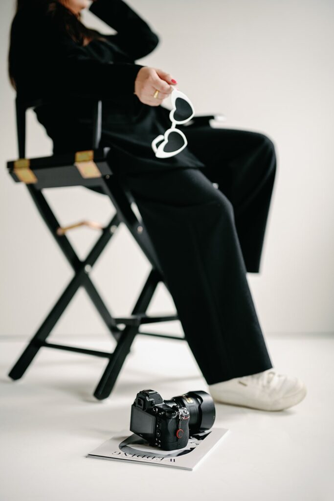 Wedding content creator wearing a black pant suit and holding white heart sunglasses while sitting in a director's chair in a studio