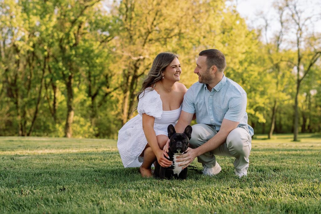 Couple crouches down to pet their black Frenchie during their engagement photo session in Indianpolis