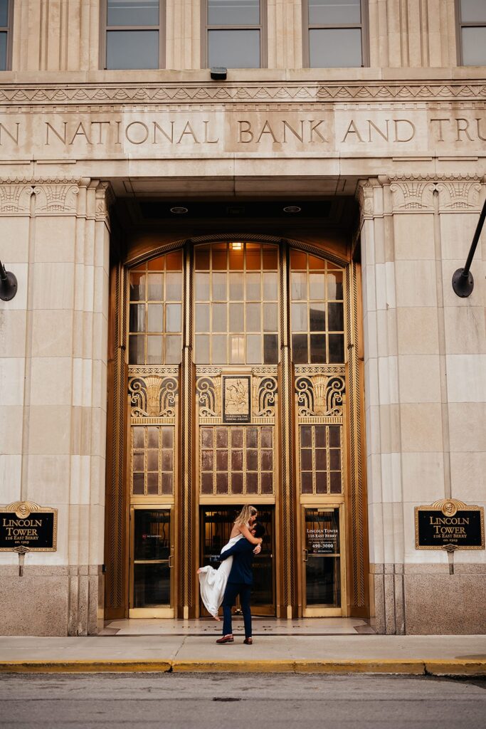 Groom lifts bride into the air in front of the gold doors of the Indianapolis National Bank