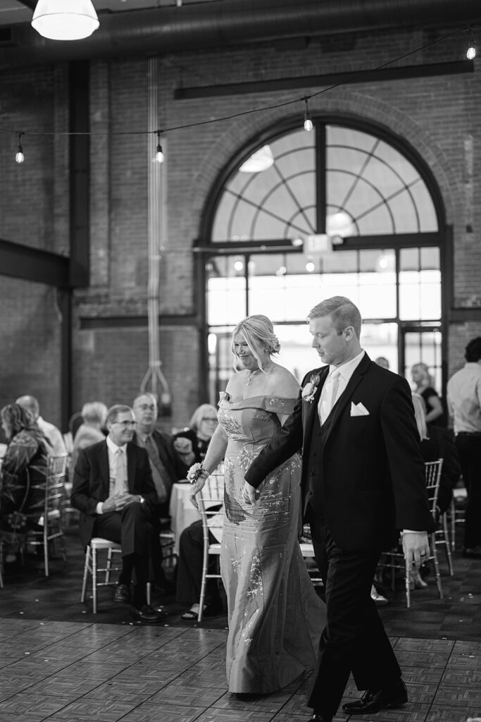 Black and white photo of a groom and his mother walking out onto the dance floor