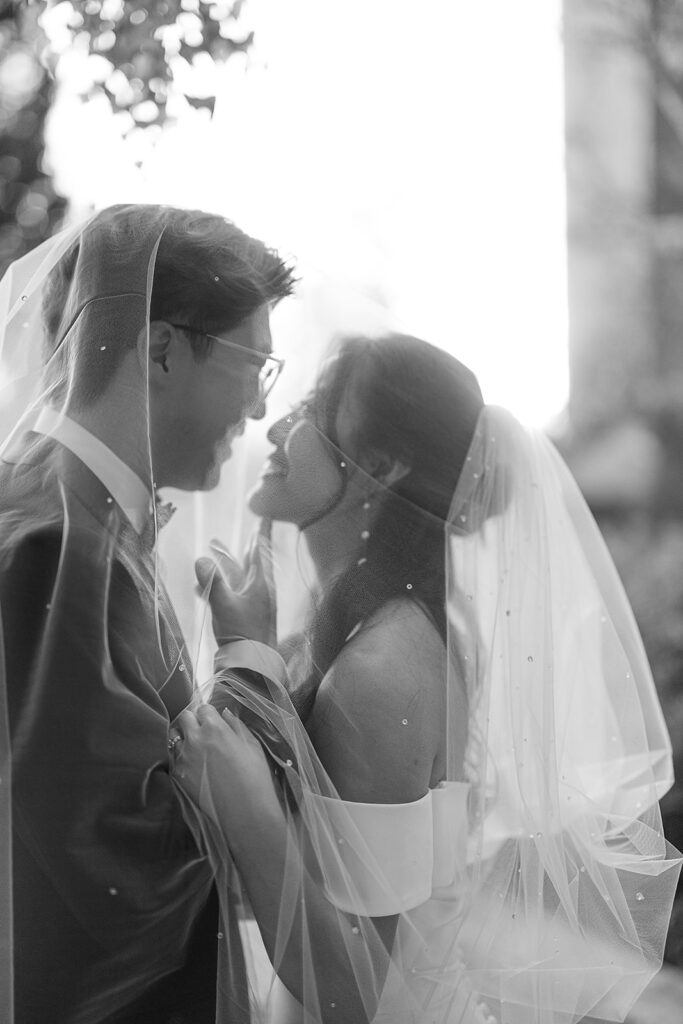 Black and white wedding photo of a bride and groom smiling under her veil