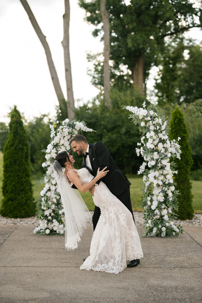 Groom dipping the bride in front of a floral arch outdoors, both laughing.