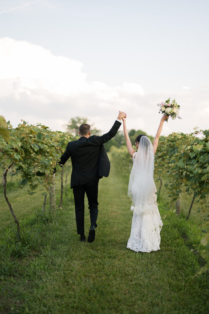 Bride and groom walking hand in hand through vineyard rows, bride holding bouquet.