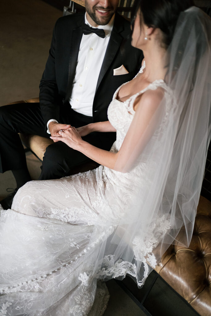 Close-up of bride and groom holding hands while seated, with the bride’s lace dress and veil in focus.