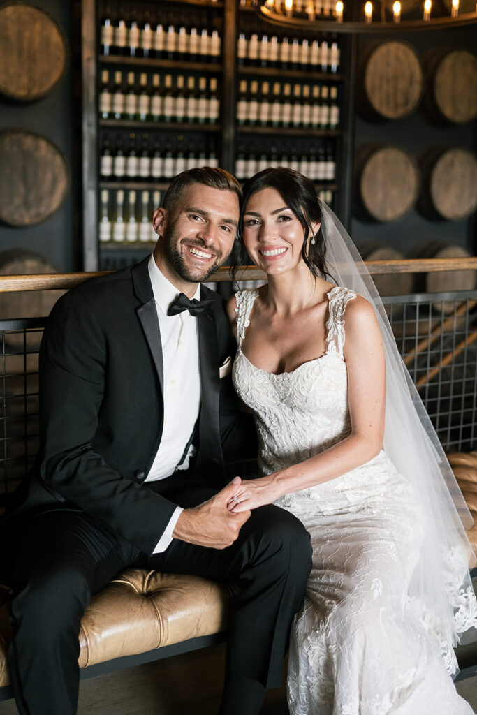 Smiling bride and groom seated closely, holding hands in front of stacked wine bottles and barrels.