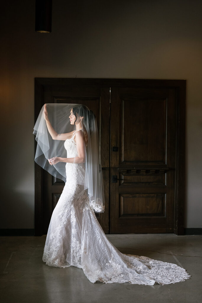 Bride standing alone in front of wooden doors, holding her veil with soft light highlighting her lace gown.