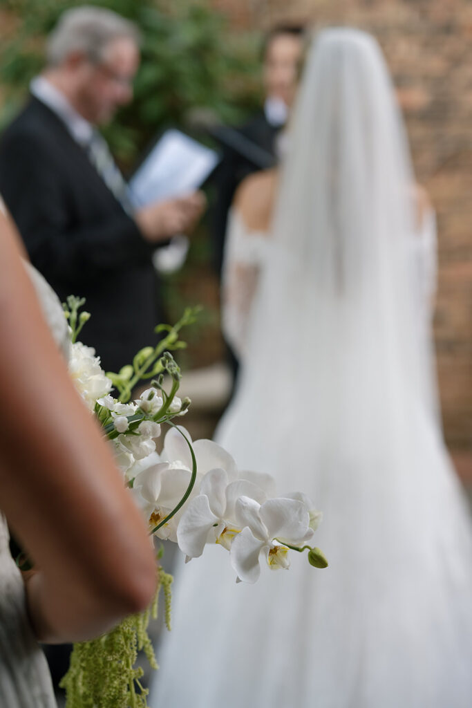 Close-up of bridesmaid’s orchid bouquet with bride and officiant blurred in the background.