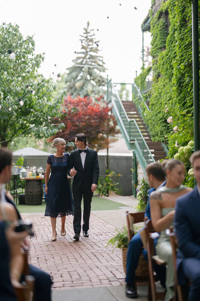 Groom walking down the aisle with his mom, surrounded by guests.