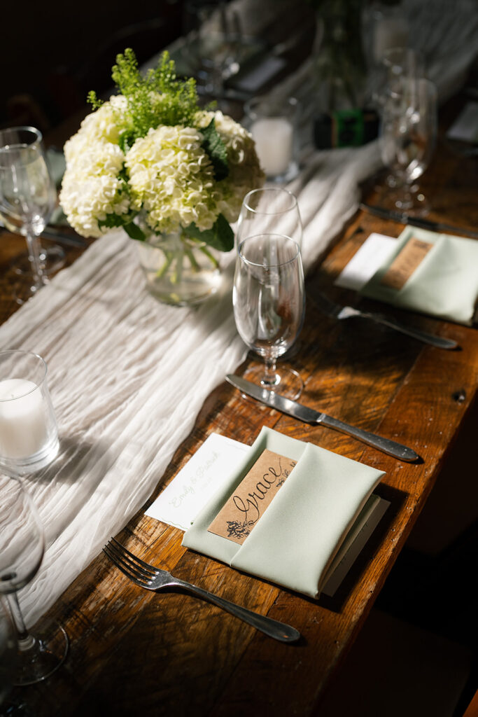ooden reception table with white hydrangea centerpiece, candles, and handwritten place card tucked into a sage napkin.
