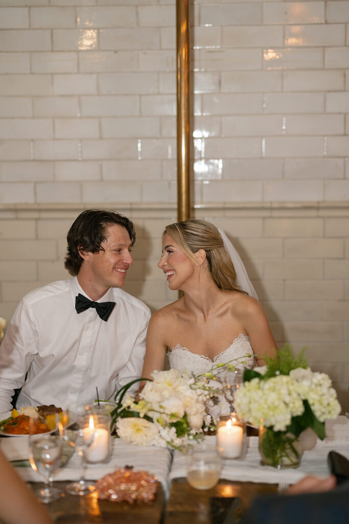 Bride and groom smiling at each other at their sweetheart table, surrounded by florals and candlelight.
