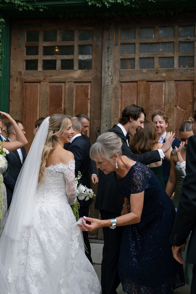 Bride smiling while someone adjusts her dress as guests celebrate around her.