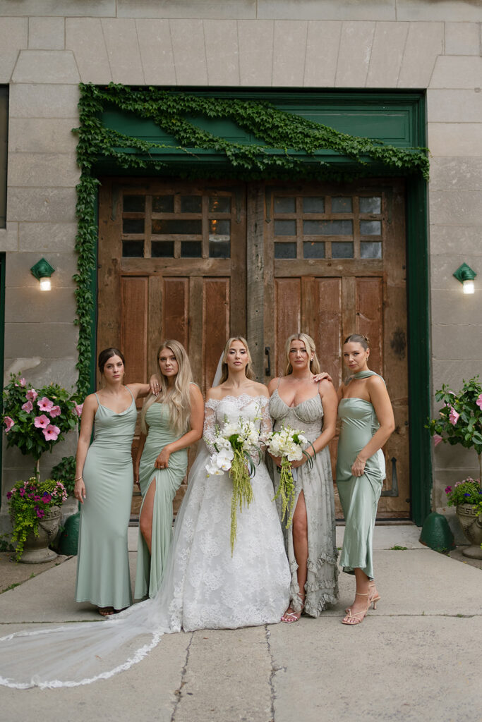 Bride with bridesmaids in sage green dresses posing in front of rustic wood doors at Firehouse Chicago.